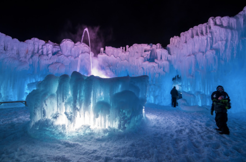Colorful ice formations illuminated at night, with a fountain and people exploring the winter wonderland.