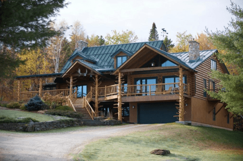 A large log cabin with a green metal roof, surrounded by trees and a gravel driveway, set in a scenic landscape.