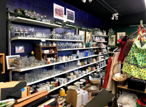 A cluttered thrift store shelf filled with various glassware, dishes, and decorative items against a blue wall.
