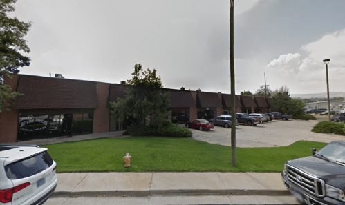A brick commercial building with parked cars and green grass in front, under a cloudy sky.