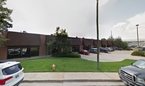 A row of brick commercial buildings with parked cars and green grass in front, under a cloudy sky.