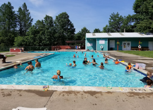 A busy outdoor pool with people swimming and playing, surrounded by trees and a building in the background.