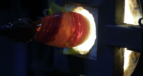 A close-up of a glassblower shaping a vibrant red and orange glass piece in a glowing furnace.