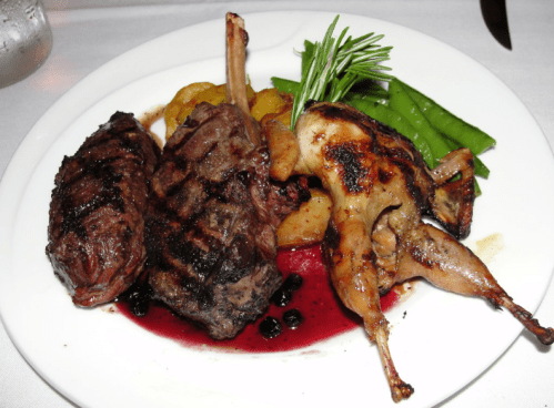 Grilled meats including lamb chops and quail, served with green beans and garnished with rosemary on a white plate.