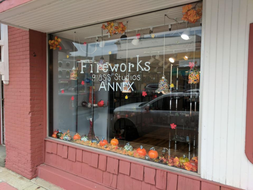 A storefront window displays "Fireworks Glass Studios Annex" with autumn decorations, including pumpkins and colorful leaves.