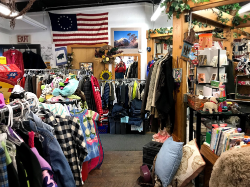 A cozy thrift store filled with clothing racks, books, and decor, featuring a vintage American flag on the wall.
