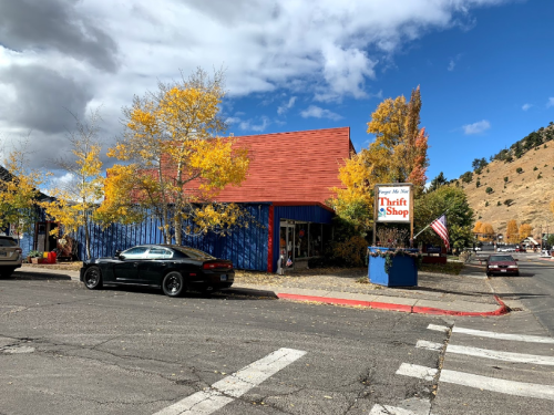 A colorful thrift shop with autumn trees and a blue sky, located on a street with a parked car and an American flag.