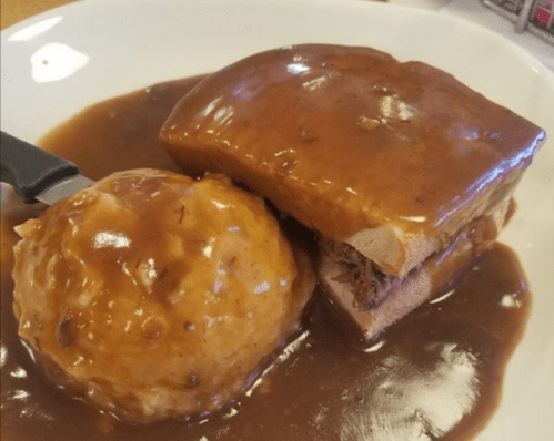 A plate featuring a slice of meatloaf covered in gravy, alongside a scoop of mashed potatoes also topped with gravy.