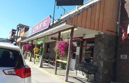 A colorful café with flower planters, benches, and a green mascot outside, set in a charming street scene.