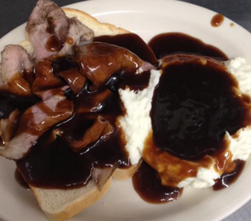 Plate of sliced roast beef on bread, topped with brown gravy, served alongside mashed potatoes with additional gravy.