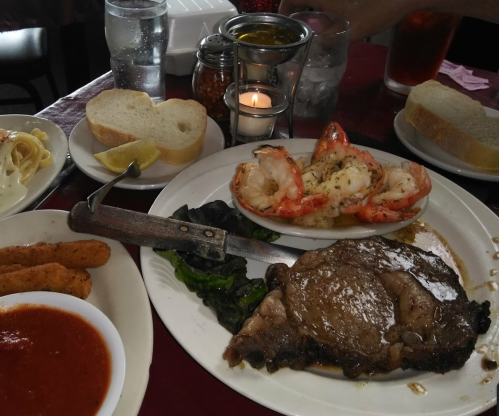 A plate featuring a steak, shrimp, bread, and sides, with drinks and condiments in the background.