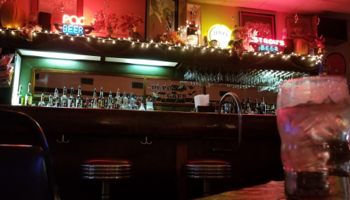 A dimly lit bar with a wooden counter, shelves of bottles, and neon signs, creating a cozy atmosphere.