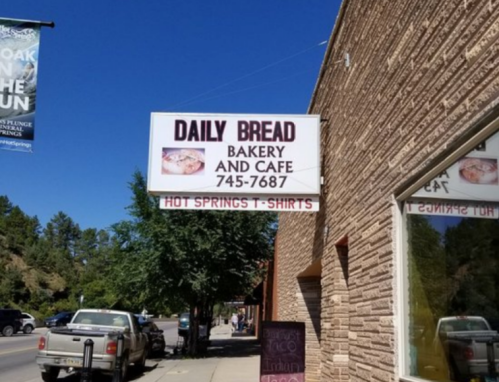 Sign for "Daily Bread Bakery and Cafe" with phone number, located in a tree-lined street. Hot Springs T-shirts below.
