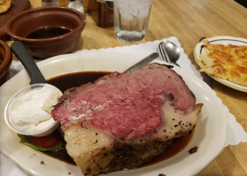 A large slice of roast beef served with a side of dipping sauce and a dollop of cream, alongside a plate of cheesy potatoes.