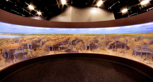 Panoramic view of a prehistoric village with round huts, surrounded by a landscape of fields and water.