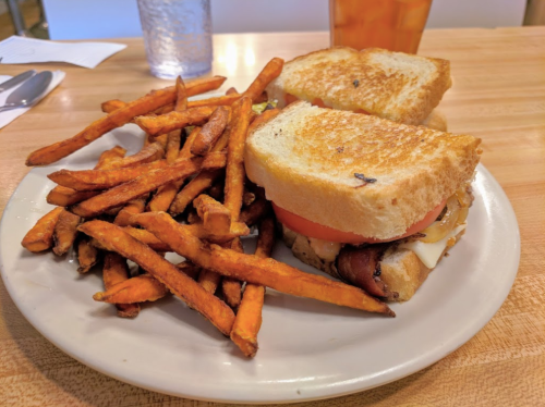A plate with a sandwich made of toasted bread, bacon, cheese, and tomato, served with a side of sweet potato fries.