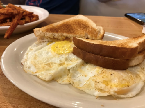A plate with two fried eggs and two slices of toasted bread, with a side of sweet potato fries in the background.