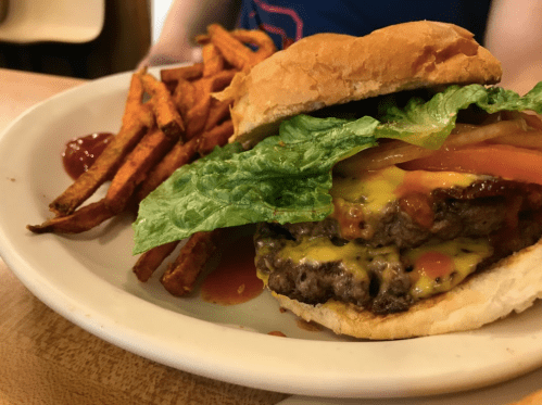 A close-up of a cheeseburger with lettuce, tomato, and ketchup, served with a side of sweet potato fries.