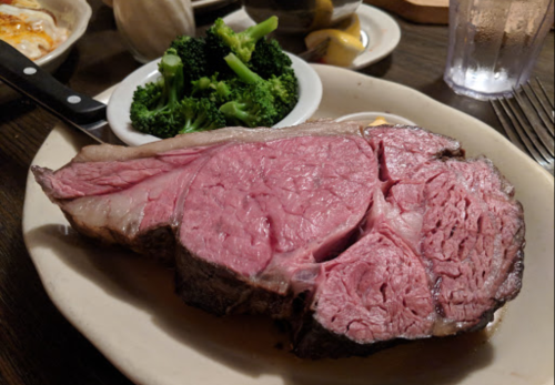 A large slice of medium-rare prime rib served with broccoli and sides on a plate.