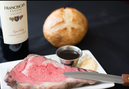 A plate with a slice of prime rib, a small bowl of sauce, mashed potatoes, a roll, and a bottle of Cabernet Sauvignon.