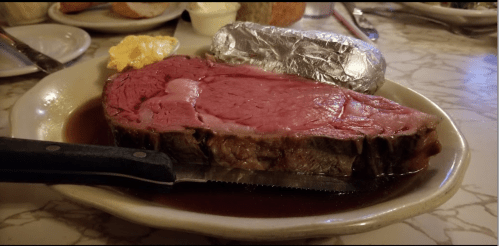 A plate with a large slice of rare prime rib, a baked potato wrapped in foil, and a pat of butter.