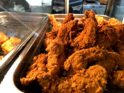 A tray of crispy, golden-brown fried chicken pieces, with some fried snacks in the background.