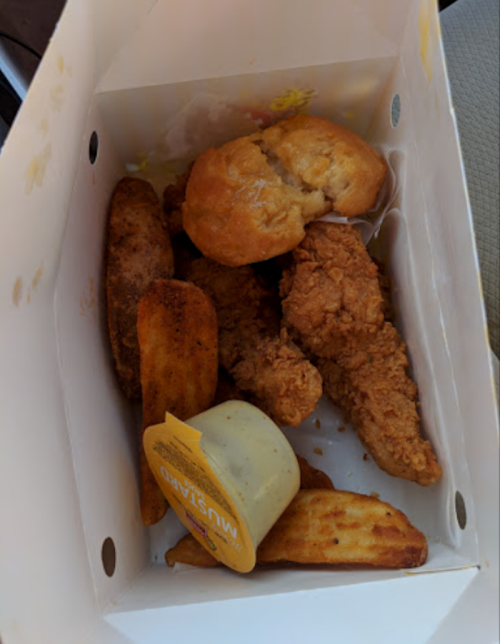 A takeout box filled with fried chicken, potato wedges, a biscuit, and a container of mustard sauce.