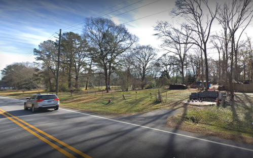 A quiet rural road with a car passing by, surrounded by trees and open fields under a clear sky.
