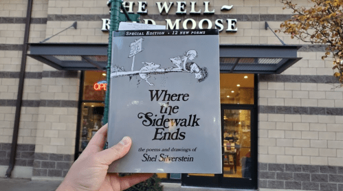 A person holds the book "Where the Sidewalk Ends" by Shel Silverstein in front of a bookstore named "The Well-Read Moose."