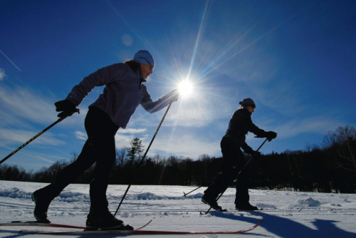 Two people cross-country skiing on a snowy landscape under a bright sun and blue sky.
