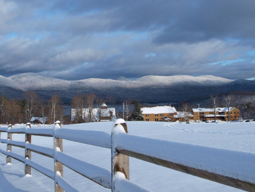 A snowy landscape with a wooden fence, mountains in the background, and a cozy building nestled in the snow.