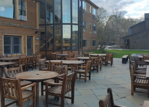 Outdoor patio with wooden tables and chairs, surrounded by buildings and trees under a cloudy sky.