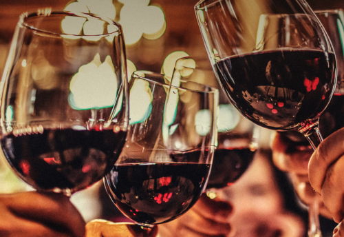 A close-up of several hands holding wine glasses, toasting with red wine against a blurred, warm background.