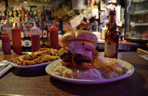 A hearty burger with toppings, fries, and a bottle of Honey Brown beer on a bar counter with a lively background.