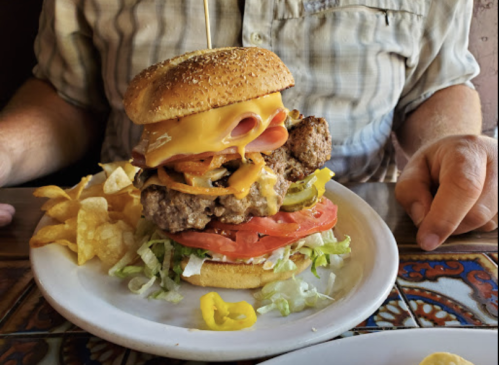 A towering burger with cheese, ham, lettuce, tomato, pickles, and jalapeños, served with a side of chips.