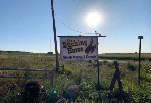 Sign for "The Bucking Horse Steak House & Lounge" in a rural setting with a clear blue sky and sun.