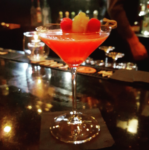 A vibrant red cocktail garnished with raspberries and a slice of fruit, served in a elegant martini glass on a bar counter.