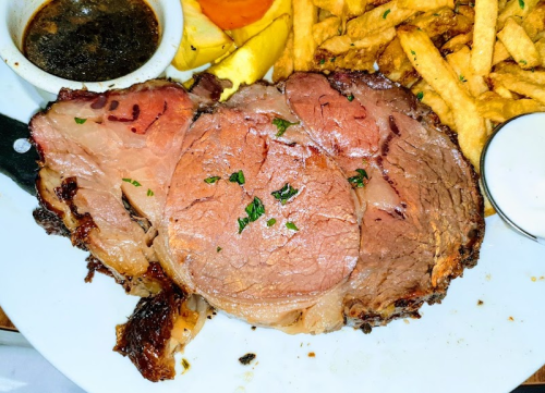 A plate featuring a thick slice of prime rib, garnished with parsley, alongside fries and dipping sauces.