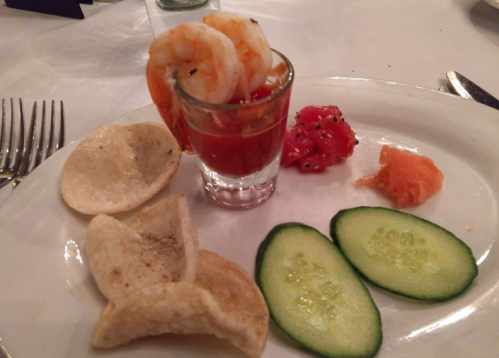 A plate with shrimp cocktail in a shot glass, cucumber slices, smoked salmon, watermelon, and crispy snacks.