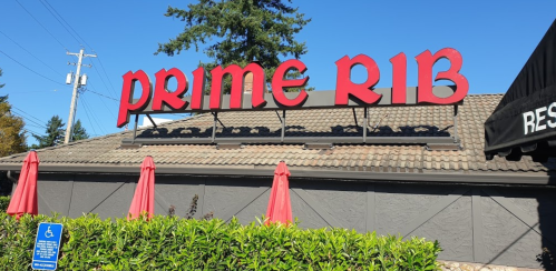 Sign for "Prime Rib" restaurant with red letters, surrounded by green bushes and red umbrellas under a clear blue sky.