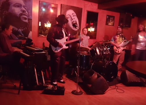 A lively band performs on stage in a dimly lit venue, with musicians playing guitar, drums, and keyboards.