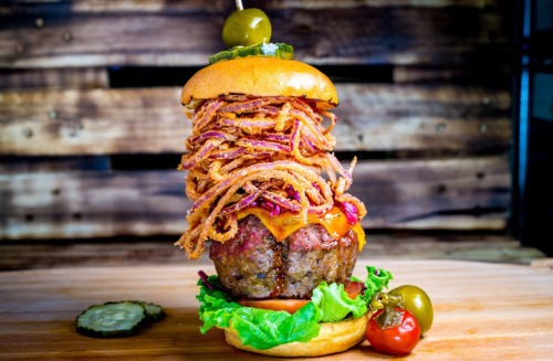 A towering burger with a beef patty, cheese, crispy onions, lettuce, and pickles on a wooden background.