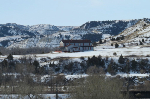 A large house on a snowy landscape, surrounded by hills and trees under a clear blue sky.