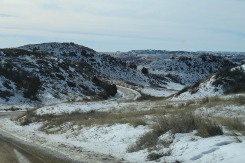 A winding road through snow-covered hills and grassy fields under a cloudy sky.