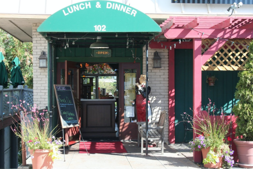A restaurant entrance with a green awning, open sign, and outdoor seating, surrounded by colorful flowers.