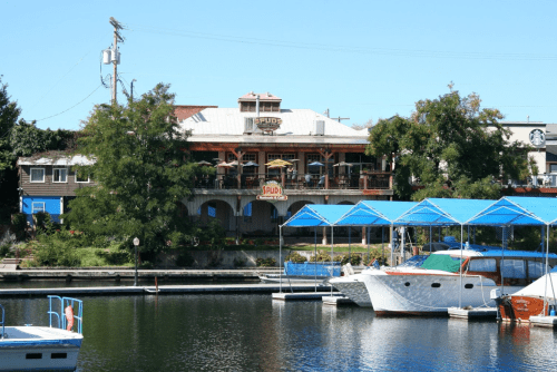 A waterfront view of a restaurant with outdoor seating, surrounded by trees and boats docked in the marina.
