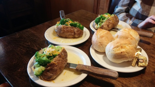 Two baked potatoes topped with broccoli and cheese, alongside three bread rolls on white plates.