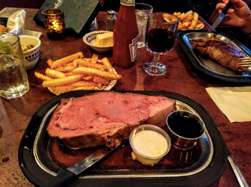 A plate with a large prime rib, dipping sauces, crinkle-cut fries, and drinks on a restaurant table.