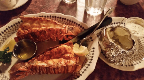 A plate with two lobster tails, a lemon wedge, a small dish of butter, and a baked potato wrapped in foil.