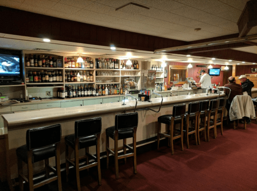 A cozy bar with a long counter, shelves of liquor, and a bartender preparing drinks. A few patrons are seated.